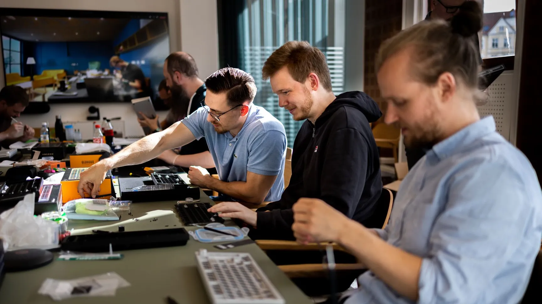 Mænd arbejdende på en samling af mekaniske tastaturer ved et bord, omgivet af værktøj og komponenter, med en stor skærm i baggrunden, der viser et moderne kontormiljø.