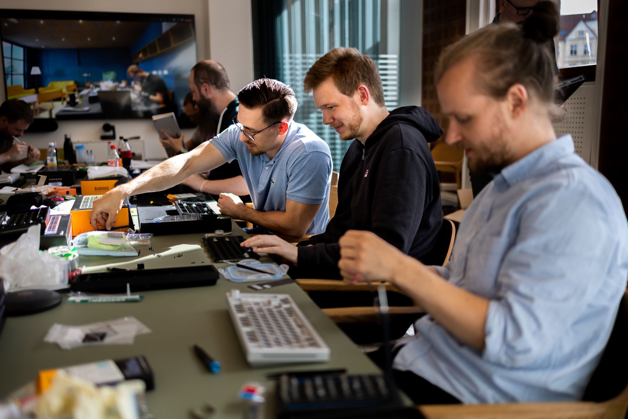 Mænd arbejdende på en samling af mekaniske tastaturer ved et bord, omgivet af værktøj og komponenter, med en stor skærm i baggrunden, der viser et moderne kontormiljø.