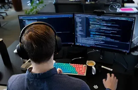 En programmør arbejder ved et skrivebord med to skærme, iført hovedtelefoner. Skærmene viser kode, og der er en farverig mekanisk tastatur og en kop kaffe foran dem.