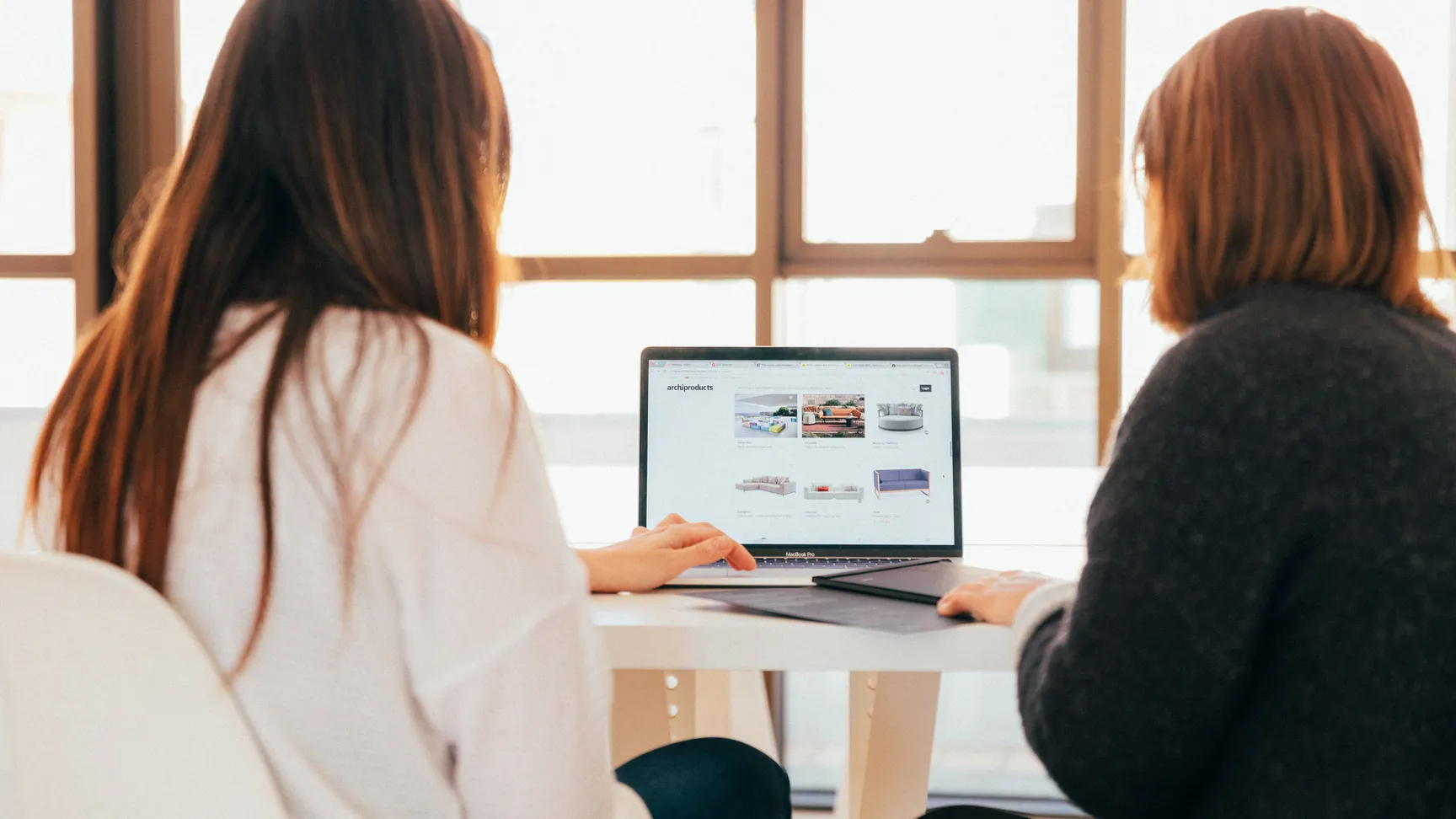 To personer sidder ved et bord og ser på en laptop, hvor der vises designmøbler på en hjemmeside. Sollys strømmer ind gennem vinduet, og der er en afslappet atmosfære i rummet.