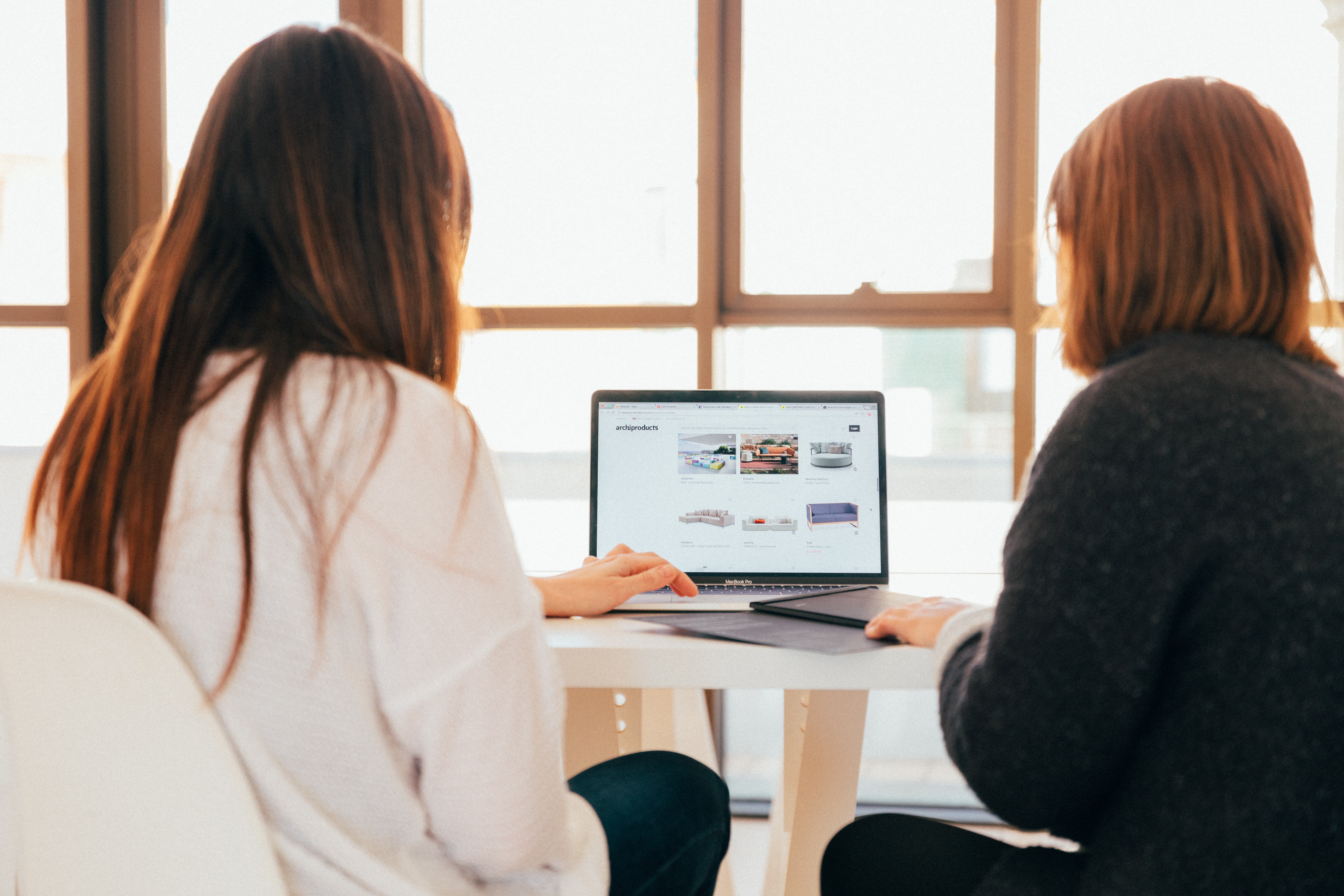 To personer sidder ved et bord og ser på en laptop, hvor der vises designmøbler på en hjemmeside. Sollys strømmer ind gennem vinduet, og der er en afslappet atmosfære i rummet.