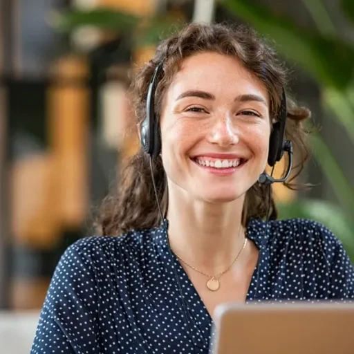 Smilende kvinde med headset, der arbejder ved en laptop i et moderne kontormiljø med grønne planter i baggrunden.