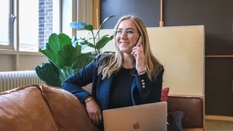 En glad kvinde sidder på en sofa og taler i telefon, mens hun arbejder på en bærbar computer i et moderne, lyst kontorlokale med planter i baggrunden.