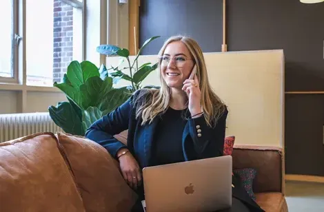En glad kvinde sidder på en sofa og taler i telefon, mens hun arbejder på en bærbar computer i et moderne, lyst kontorlokale med planter i baggrunden.