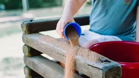 Et barn hælder sand fra en blå skål ned i en træt boks, mens der er en rød spand i nærheden; fokus på leg og kreativitet i en udendørs legeplads.