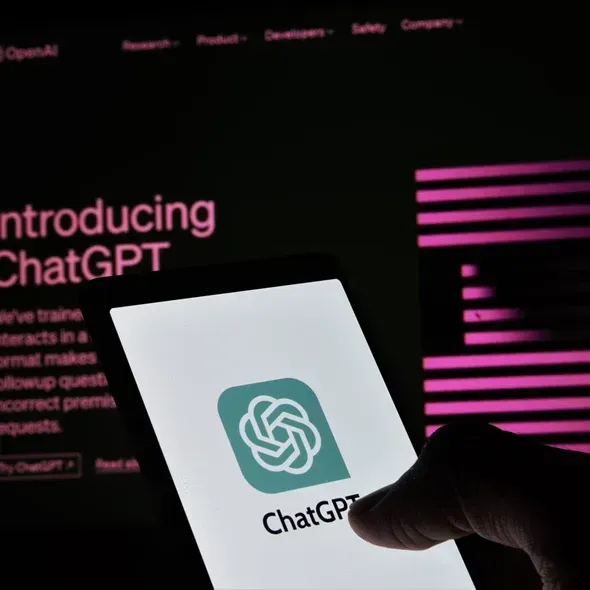 En hånd holder en smartphone, der viser ChatGPT-logoet, mens en online præsentation med teksten "Introducing ChatGPT" og en mørk baggrund kan ses i baggrunden.