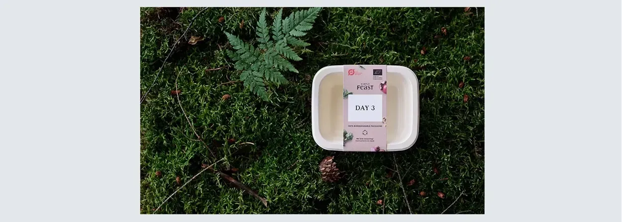 Beholder med label "DAY 3" på græsset, omgivet af enebær og bregner, der viser bæredygtig emballage.