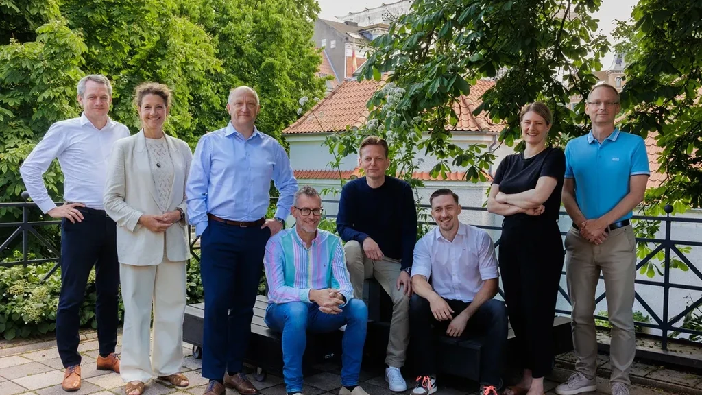 Gruppebillede af otte personer, der står og sidder i en udendørs setting med grønne træer og tagene af bygninger i baggrunden, alle iført forskellige forretningsoutfits.