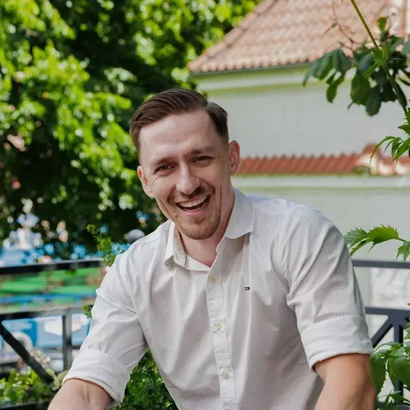 En smilende mand iført en hvid skjorte sidder på en balkon omgivet af grønne planter, med en baggrund af bygninger og træer.