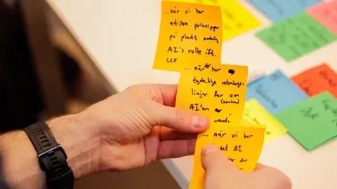 Hånd med farverige post-it sedler med håndskrevne noter, der beskriver opgaver og roller i et samarbejdsprojekt.