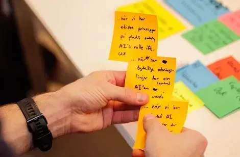 Hånd med farverige post-it sedler med håndskrevne noter, der beskriver opgaver og roller i et samarbejdsprojekt.