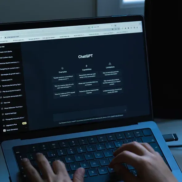 En person sidder ved en bærbar computer med en mørk skærm, der viser ChatGPTs interface med sektioner for eksempler, kapaciteter og begrænsninger.