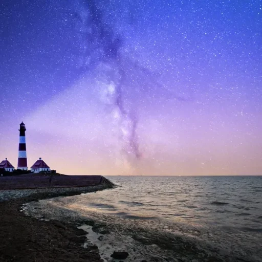 En smuk stjernehimmel over havet med en fyrtårn i forgrunden, hvor lyset fra fyret lyser op i natten, og Milky Way strækker sig over den mørkeblå himmel.