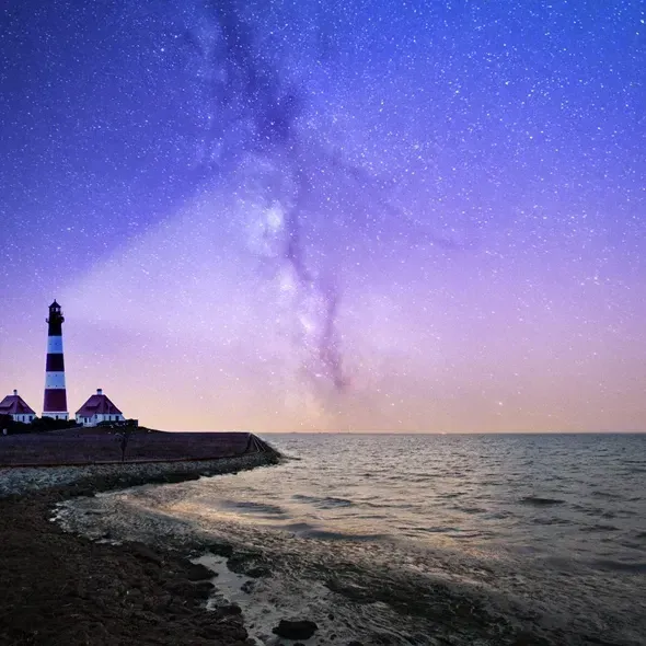 En smuk stjernehimmel over havet med en fyrtårn i forgrunden, hvor lyset fra fyret lyser op i natten, og Milky Way strækker sig over den mørkeblå himmel.