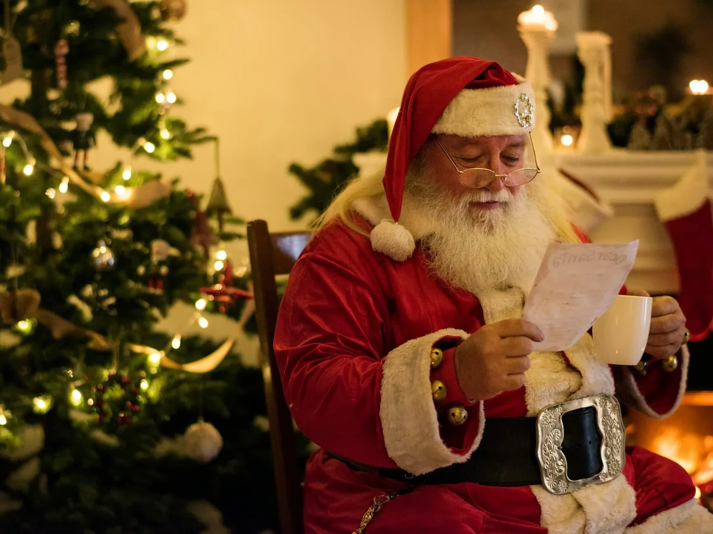 Santa Claus sitting by a cozy fireplace, reading a letter while holding a cup, with a decorated Christmas tree in the background adorned with lights and ornaments.