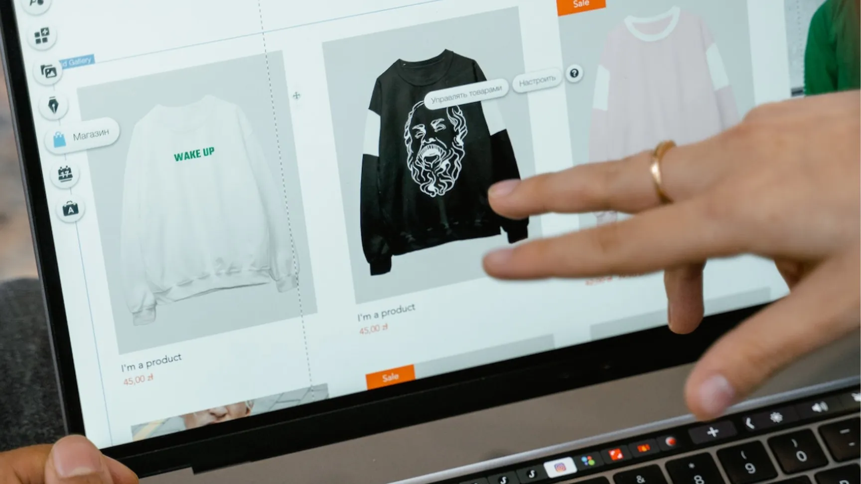Billede af en person, der interagerer med en laptop, viser en online butik med to sweatshirts: en hvid med teksten "WAKE UP" og en sort med et design af en løve.