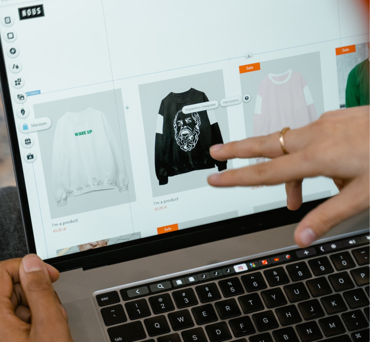 Billede af en person, der interagerer med en laptop, viser en online butik med to sweatshirts: en hvid med teksten "WAKE UP" og en sort med et design af en løve.