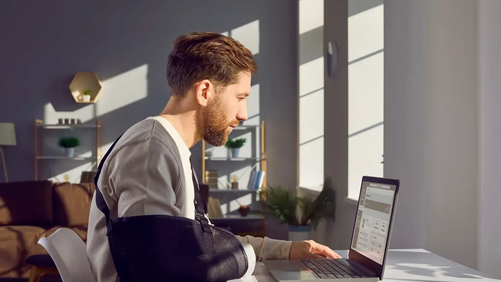 Mand med en arm i slynge arbejder ved en bærbar computer i et lyst rum med sollys, moderne indretning og planter.