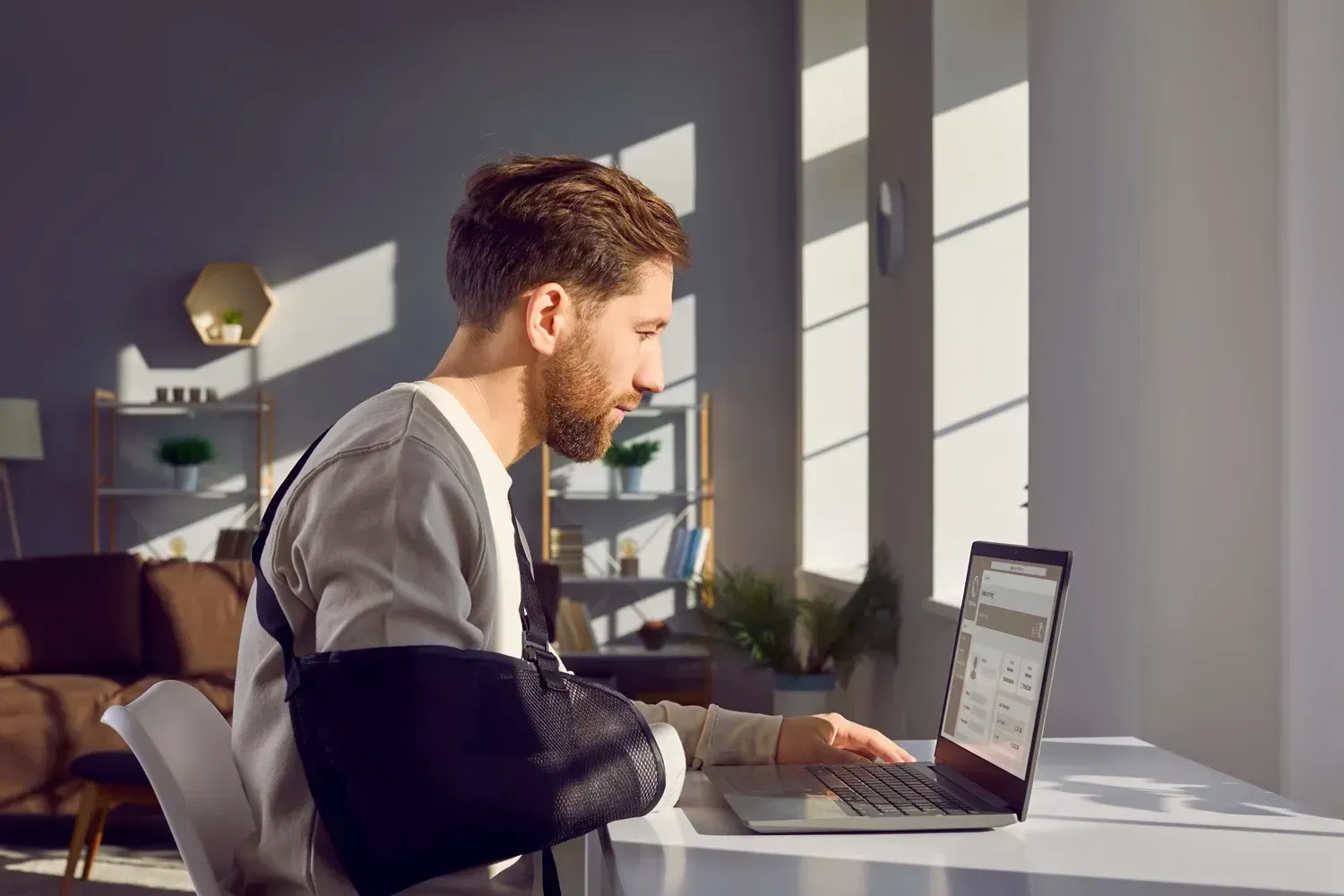 Mand med en arm i slynge arbejder ved en bærbar computer i et lyst rum med sollys, moderne indretning og planter.