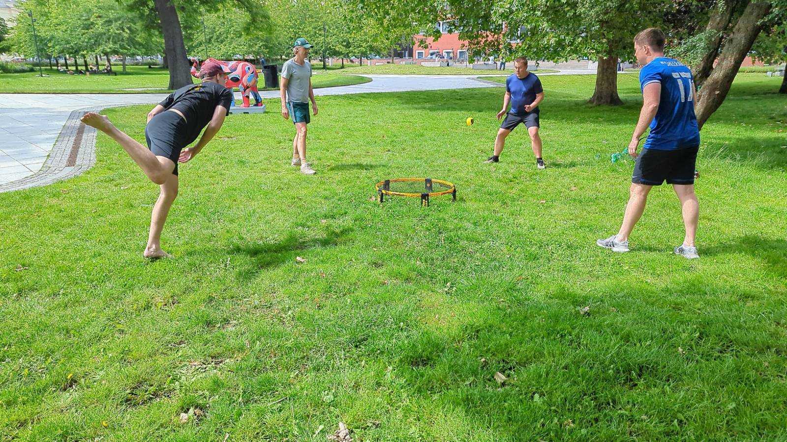 Fire personer spiller et udendørs spil med en ring på en grøn græsplæne i en park. En spiller i sort shorts står på ét ben og sparker en bold mod ringen, mens de andre spillere ser til. Det er solrigt, og der er træer i baggrunden.