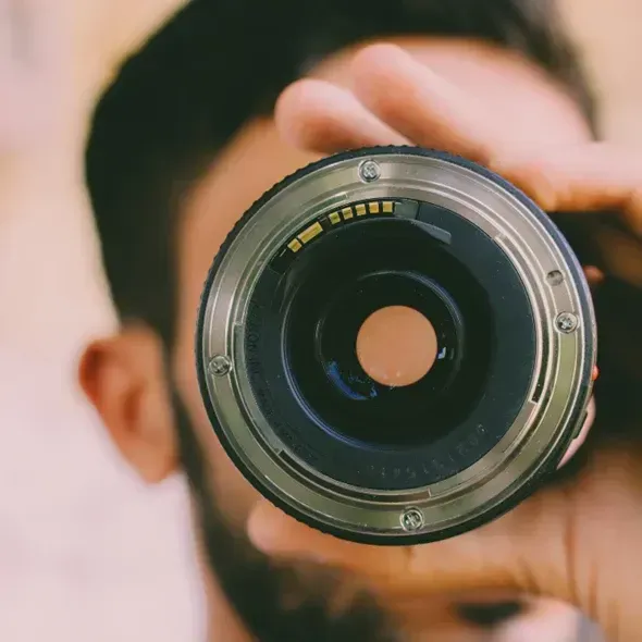 Mand holder et kameraobjektiv foran sit ansigt med en sløret baggrund.