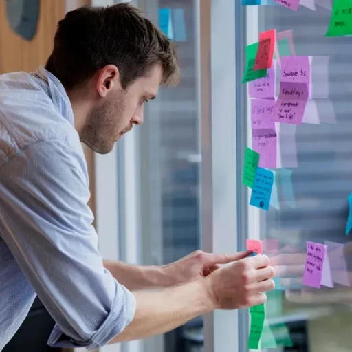 Mand står ved vinduet og sætter farverige post-its op, mens han organiserer idéer i en kreativ arbejdsindstilling.
