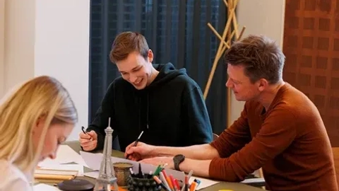 Tre personer sidder omkring et bord og tegner. To mænd, en i sort hættetrøje og en anden i brun trøje, griner og udveksler idéer, mens en kvinde i lyse hår fokuserer på sit eget papir. Der er farver, blyanter og noter spredt ud over bordet.