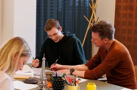 Tre personer sidder omkring et bord og tegner. To mænd, en i sort hættetrøje og en anden i brun trøje, griner og udveksler idéer, mens en kvinde i lyse hår fokuserer på sit eget papir. Der er farver, blyanter og noter spredt ud over bordet.