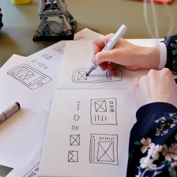 Hånd, der tegner skitser af mobilapp-interfaces med sort og blå tusch på papir, omgivet af papir med forskellige designskitser og en model af Eiffeltårnet i baggrunden.