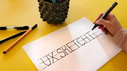 Hånd, der holder en marker og skriver "UX SKETCH" på et papir, med farverige tegneredskaber i en beholder på den gule overflade.
