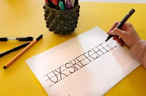 Hånd, der holder en marker og skriver "UX SKETCH" på et papir, med farverige tegneredskaber i en beholder på den gule overflade.