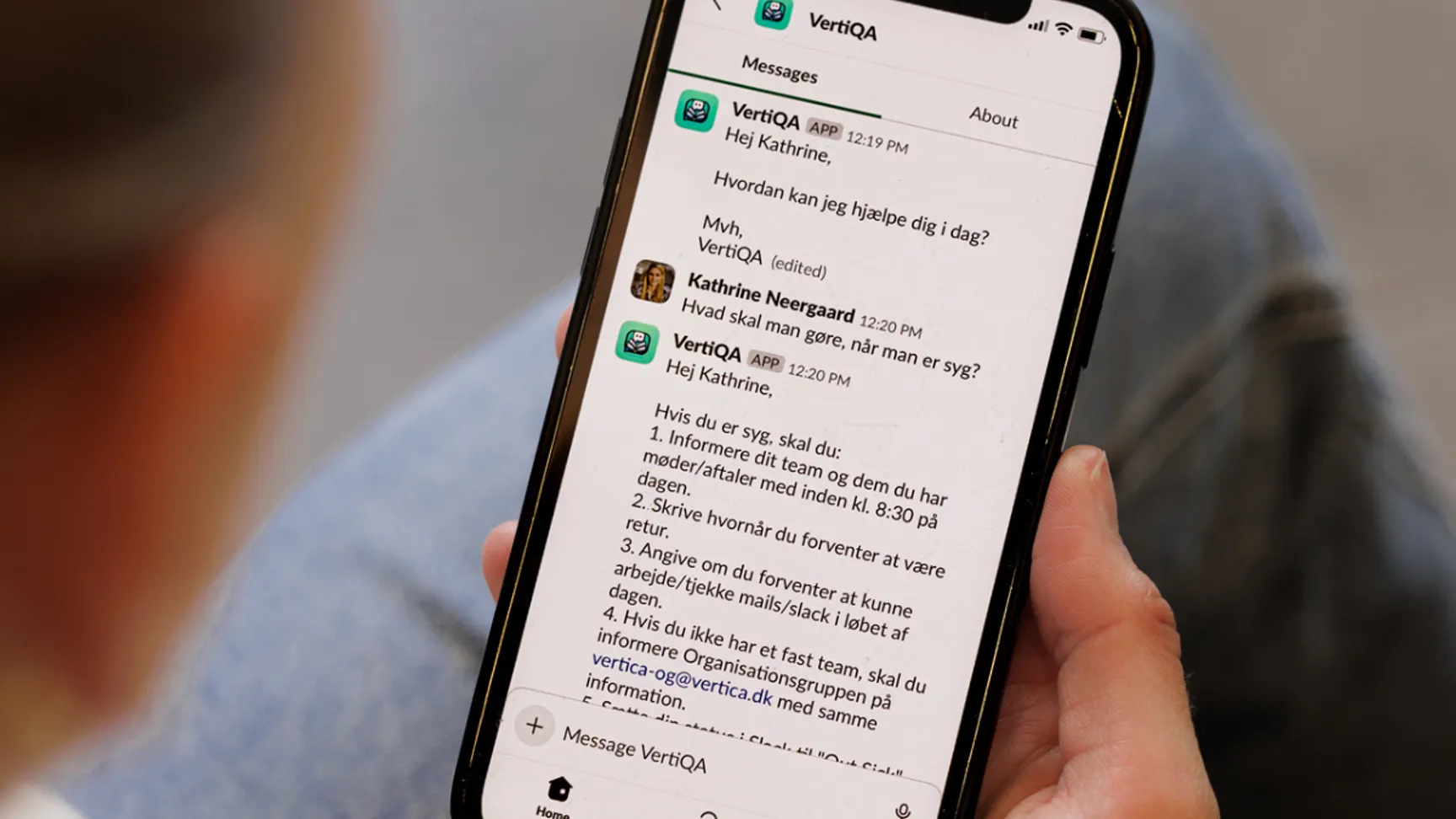 En person holder en smartphone, der viser en beskedudveksling i VertiQA-appen med en besked til "Kathrine" og svar om, hvordan man kan få hjælp. Skærmen viser detaljer om kontakten og behandlingsinstrukser.